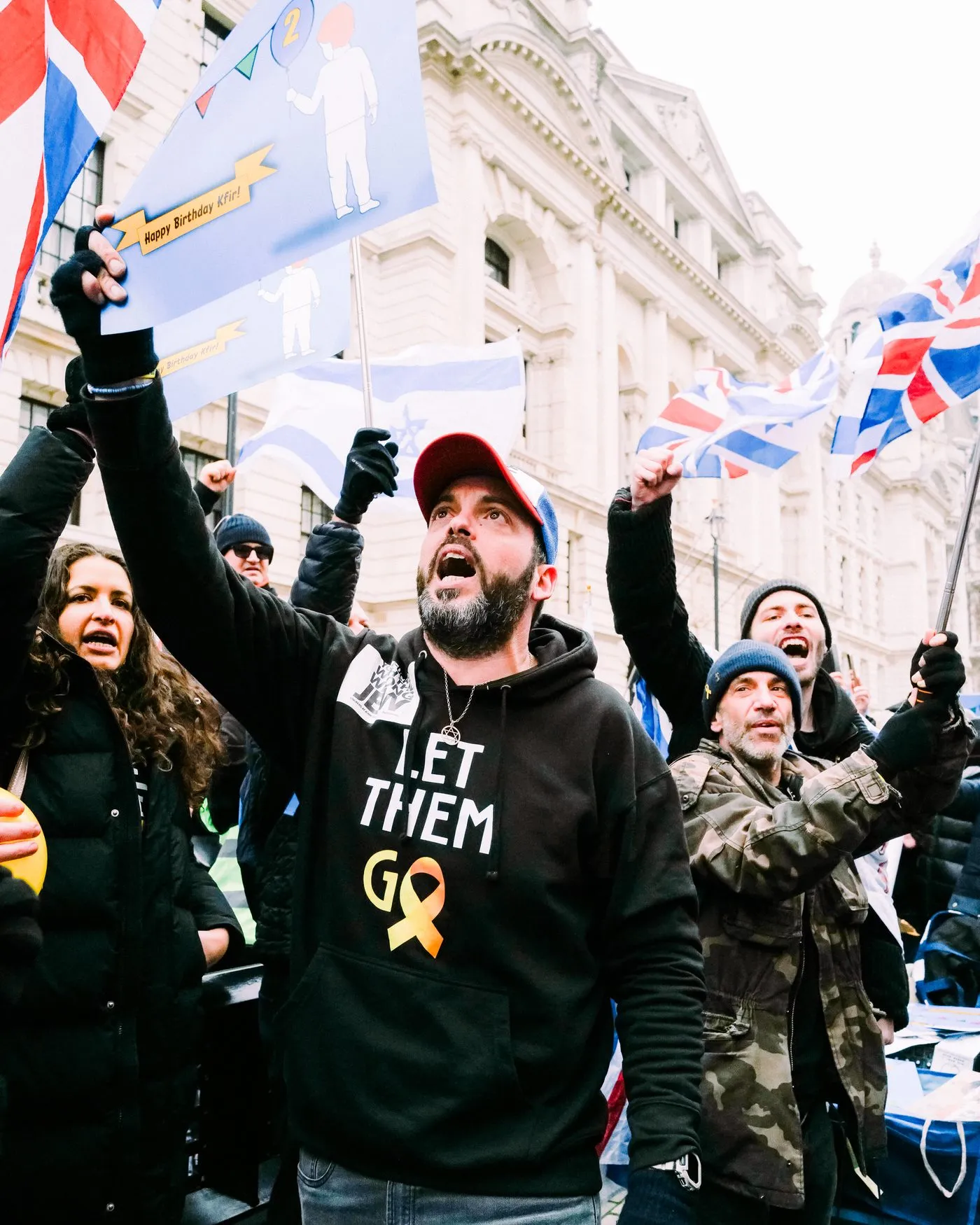 A man in a Let Them Go hoodie shouts with one arm raised outside a stone government building in Whitehall, Union flags behind him.