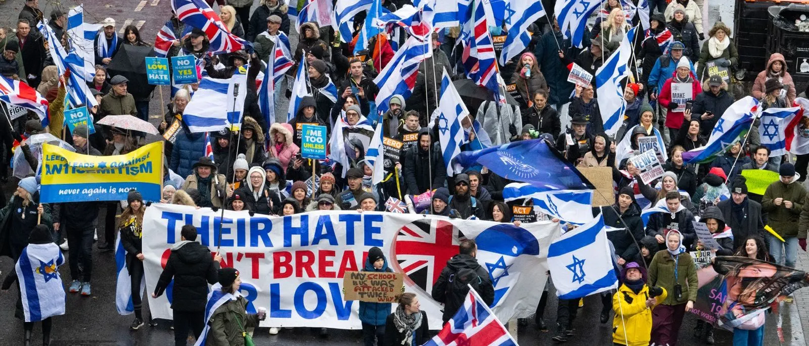 A march fills a wet London road from kerb to kerb, hundreds of people carrying Israeli and Union flags behind a banner reading Their hate will not break our love.
